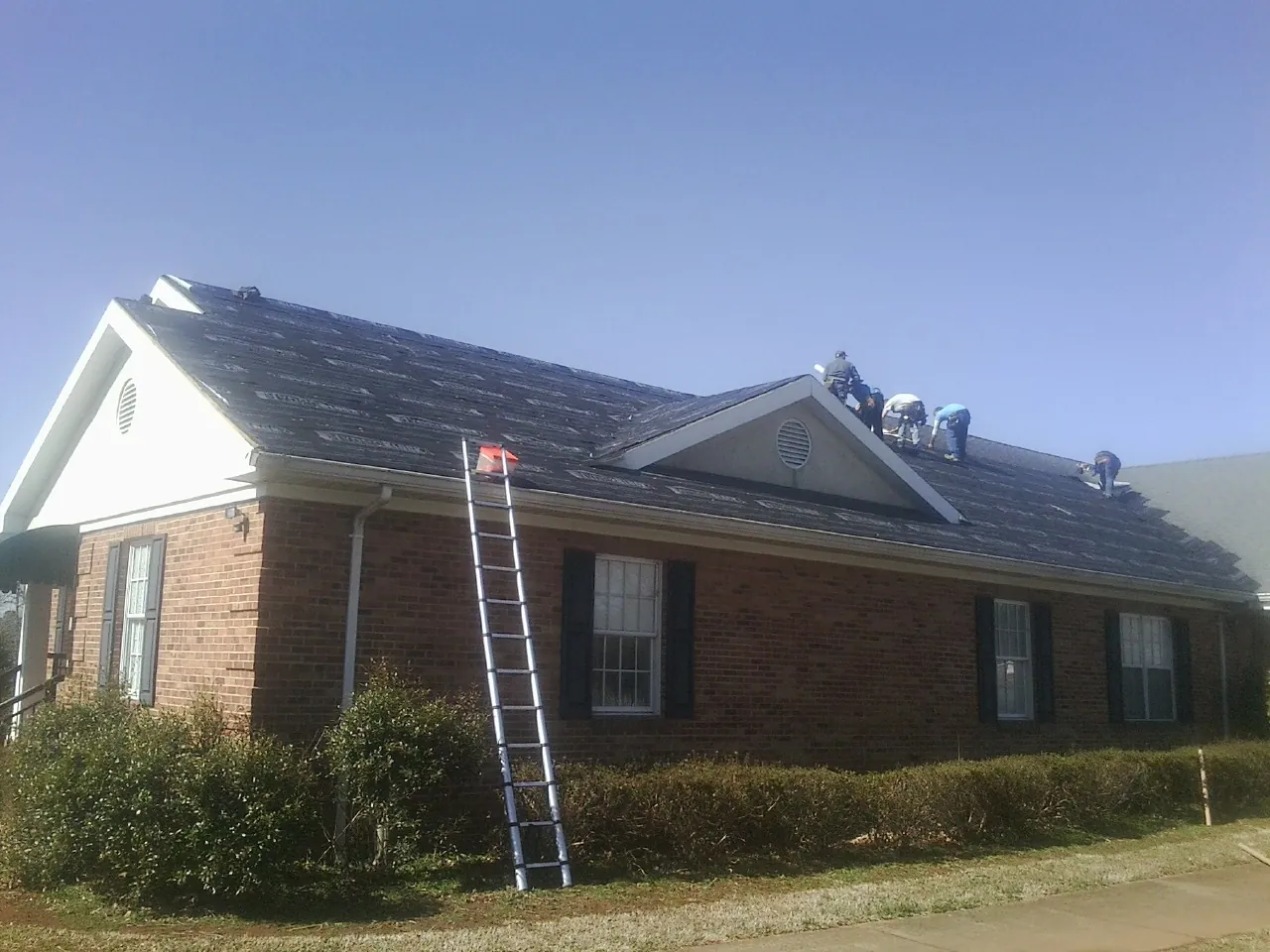 Crew tearing shingles off a brick ranch roof with a ladder set — Emergency Roof Repair work by our crew in Waukon