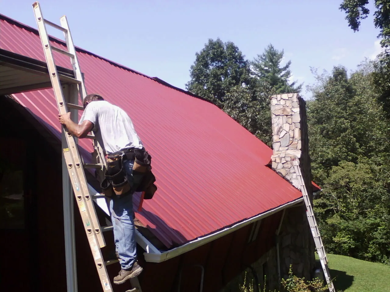 Roofer on a ladder beside a red metal roof and stone chimney — Soffit & Fascia Repair by Roofing Brothers in Waukon